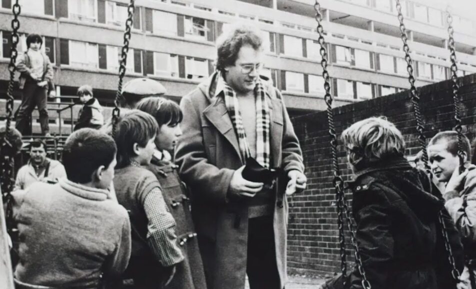 Black-and-white still from Acceptable Levels (1983): a man in a duffel coat and scarf talks with children at a playground in front of the Divis flats